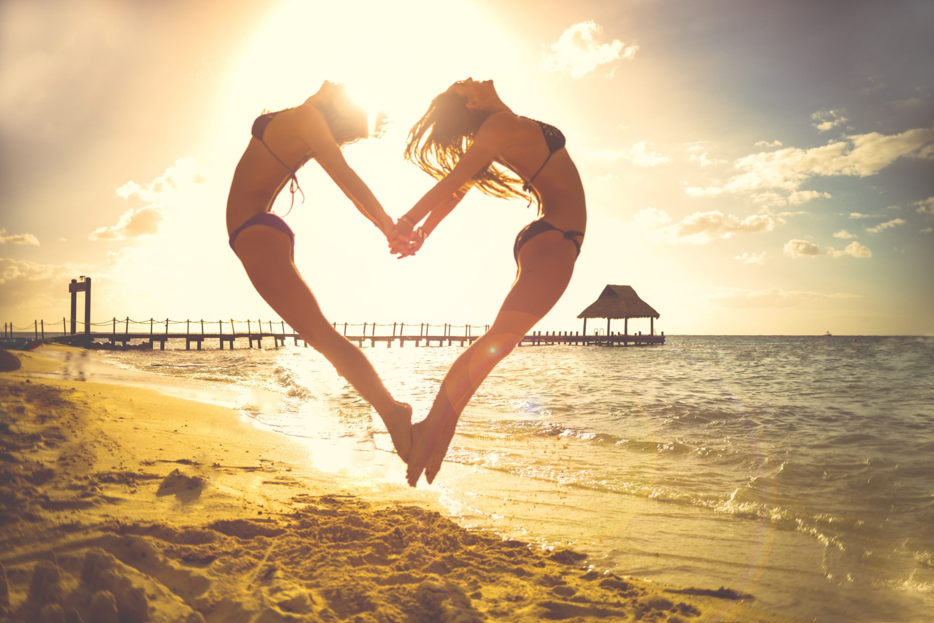 2 girls in swimwear at beach make a heart shape while jumping and holding hands